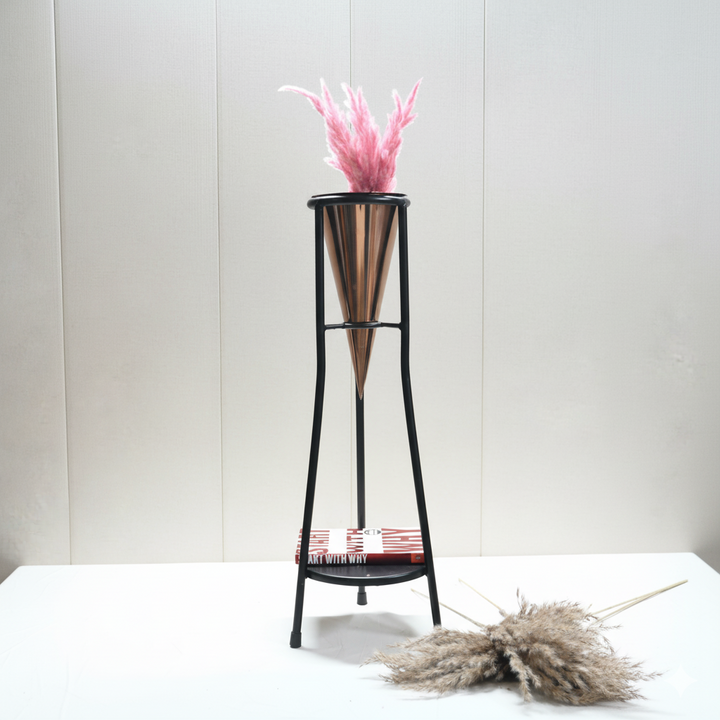 Black metal floor plant stand with a copper cone planter holding pink pampas grass, with a red and white book ("Art With Why") resting on the lower shelf.